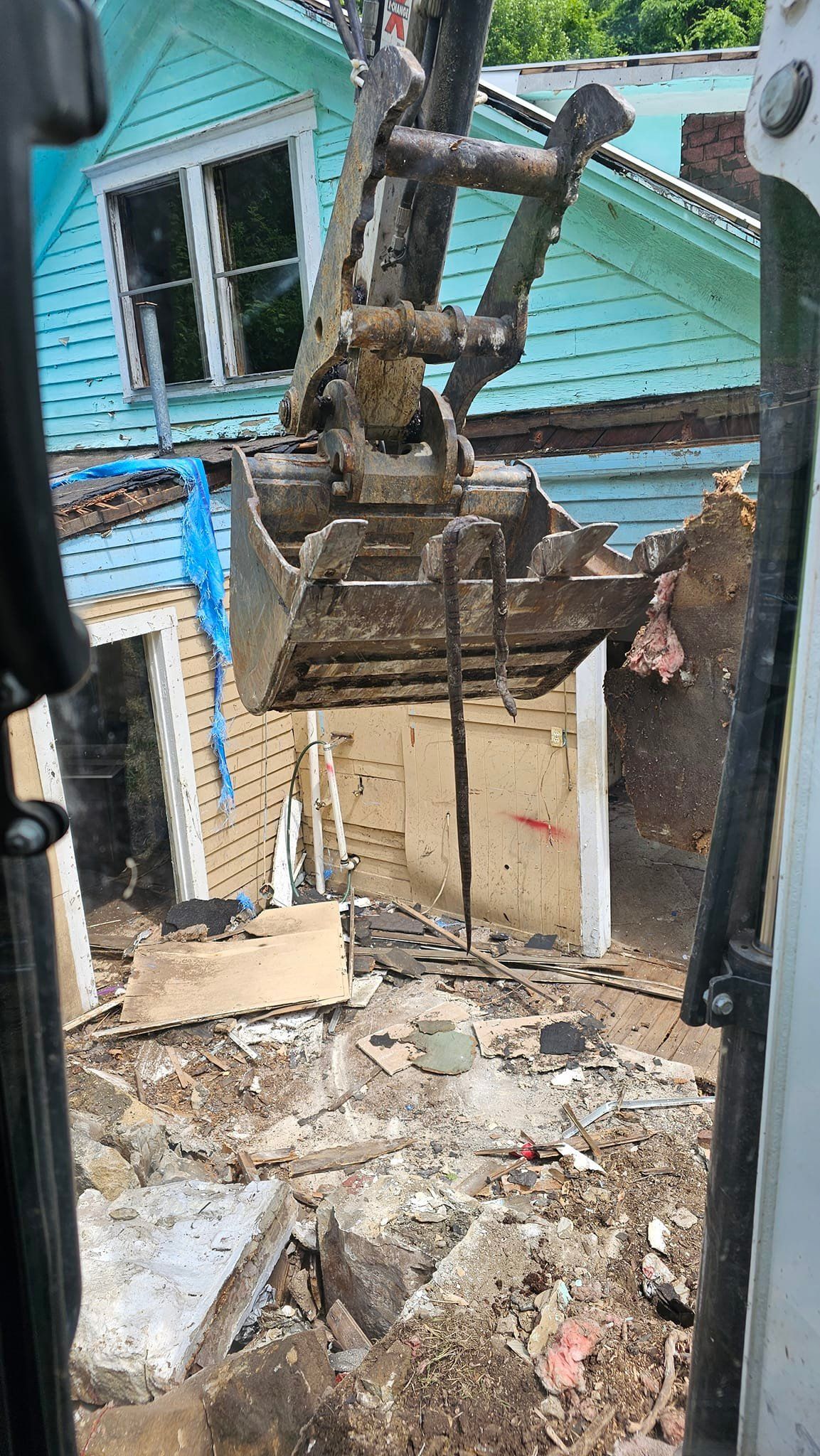 Excavator demolishing a house, showing the front wall and bucket in action. Turquoise siding and debris visible.