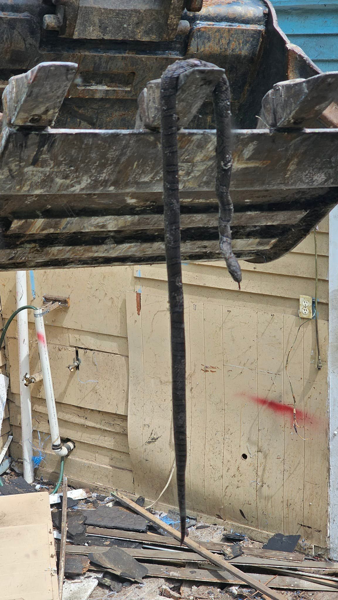 A long, dark, flexible object hangs from a construction vehicle's claw; debris visible below.