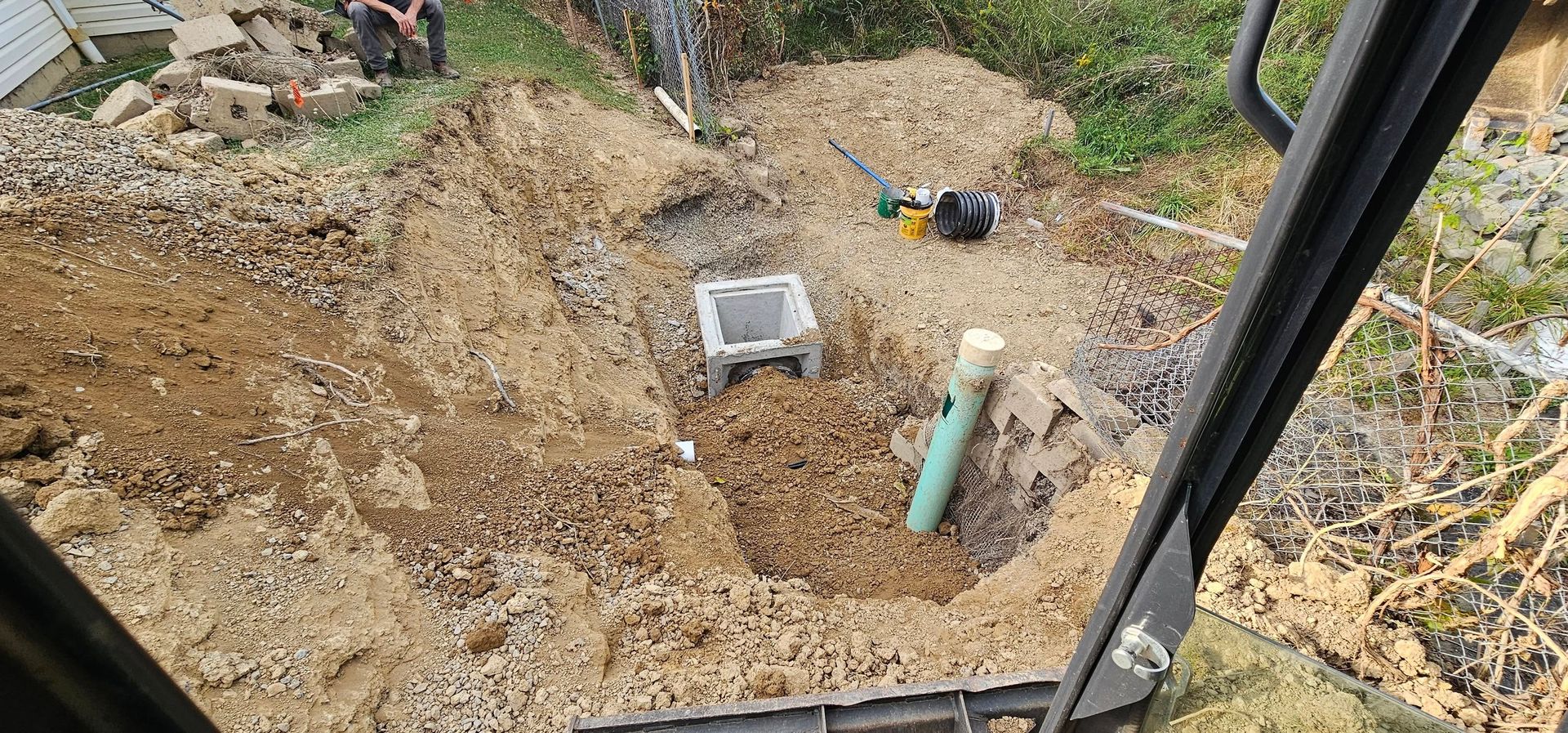 Dirt with a concrete structure and green pipe, likely sewage. From inside equipment cab.