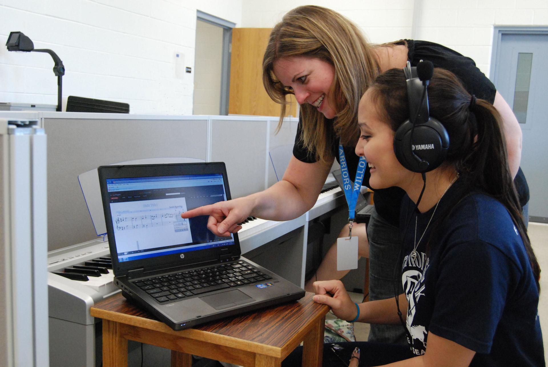 A woman is pointing at a laptop screen while another woman wearing headphones looks on.