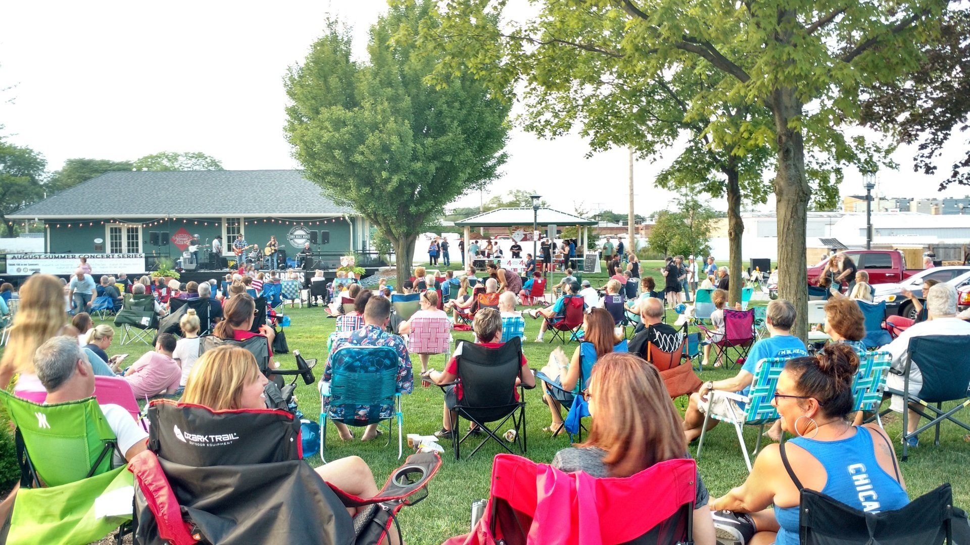 A large group of people are sitting in chairs in a park watching a concert.