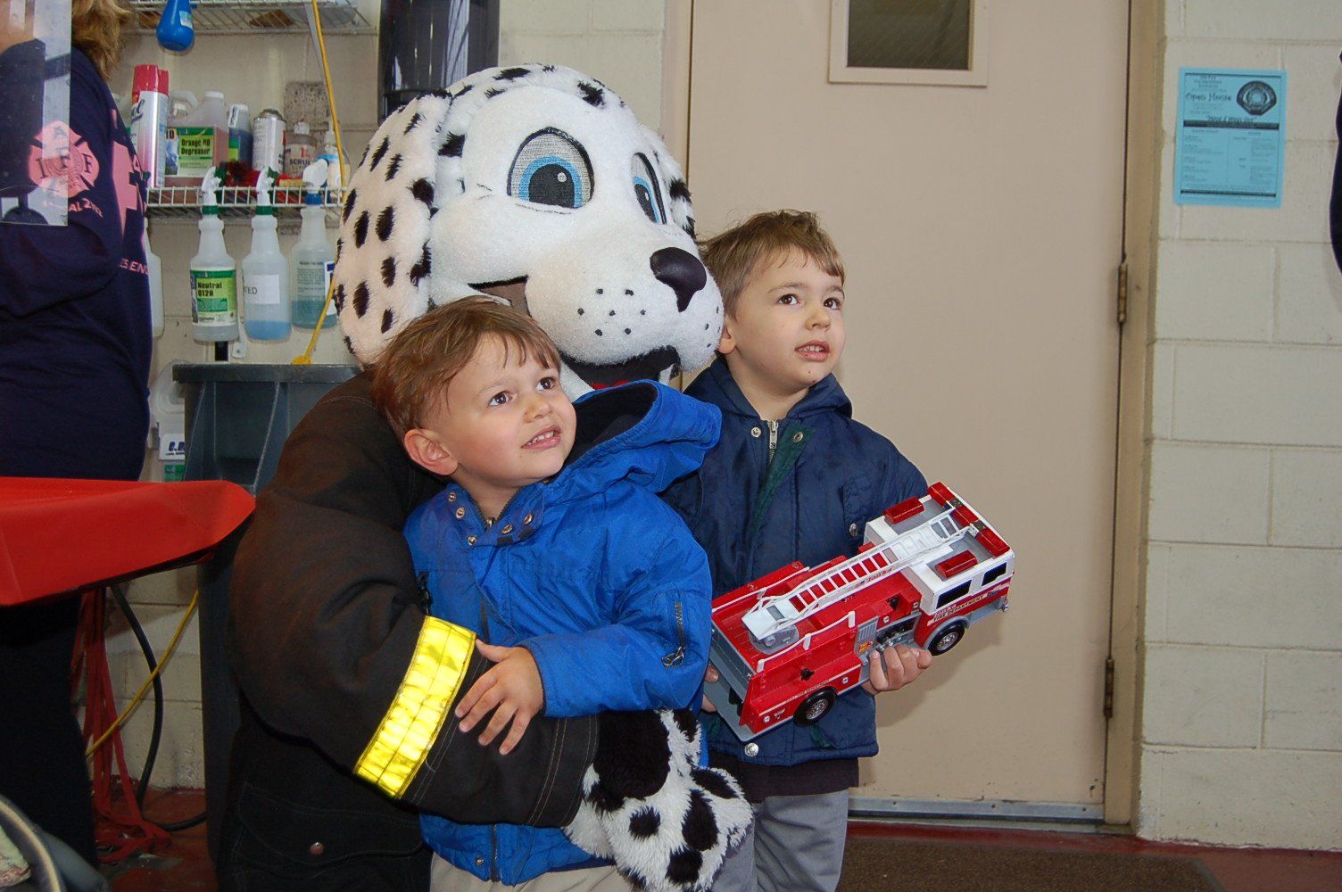 A dalmatian mascot is holding two young boys and a toy fire truck
