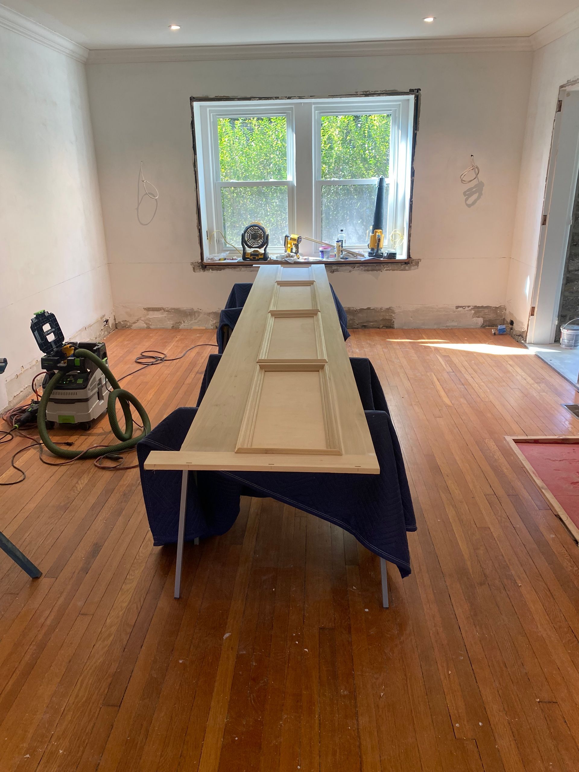 A long wooden table in a room with a window and a vacuum cleaner.