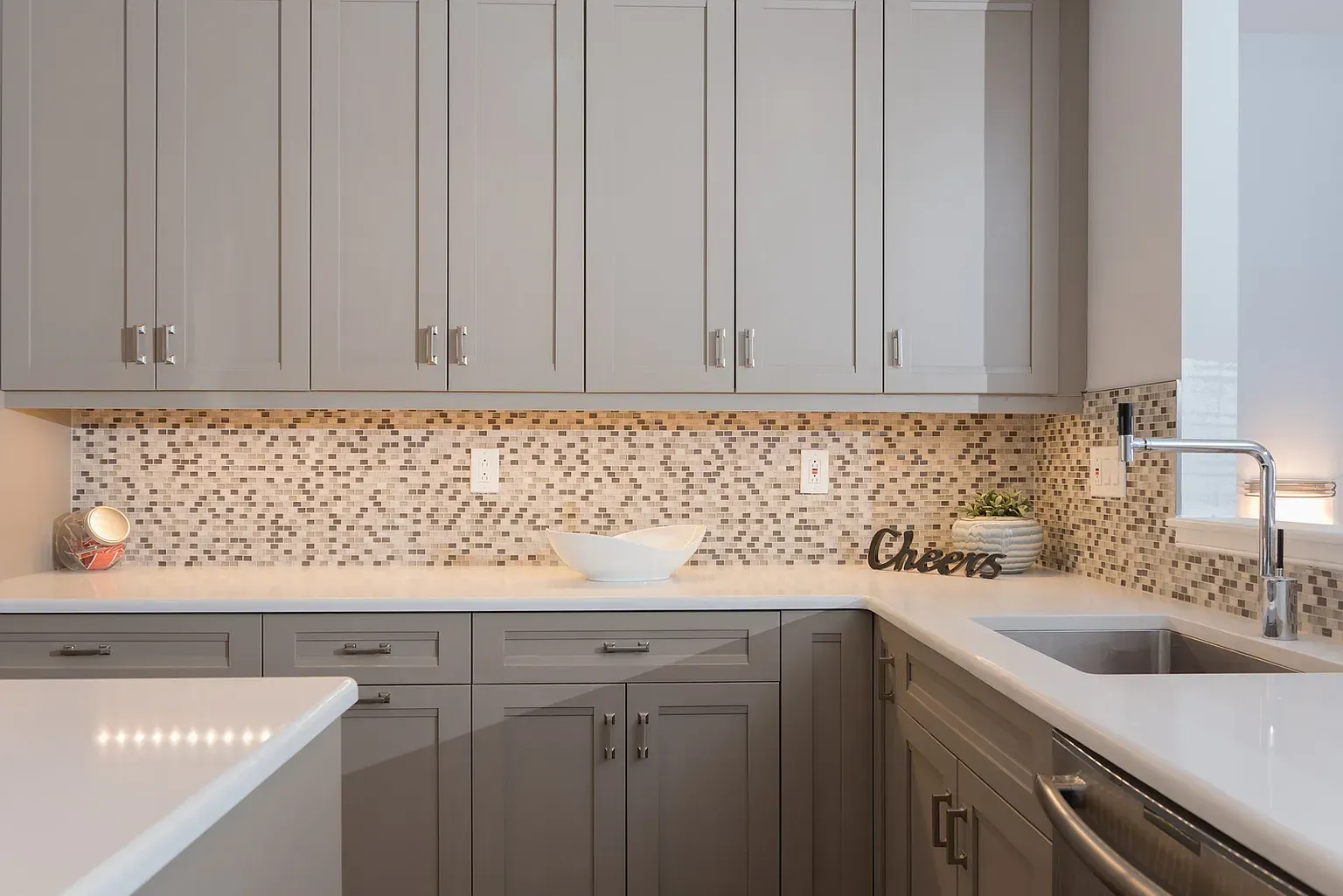 A kitchen with gray cabinets , white counter tops , a sink and a sign that says cheers.