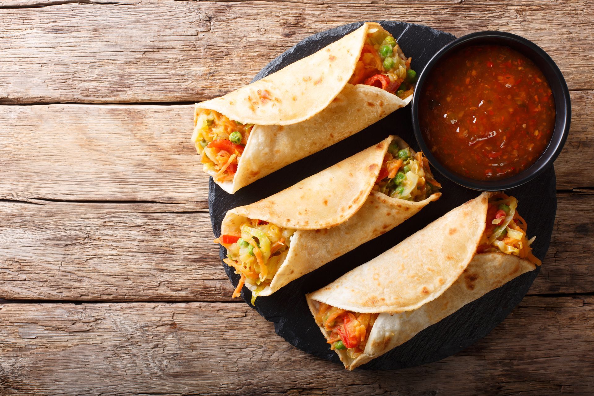 Tacos with filling and a bowl of red salsa on a black plate, set on a wooden surface.