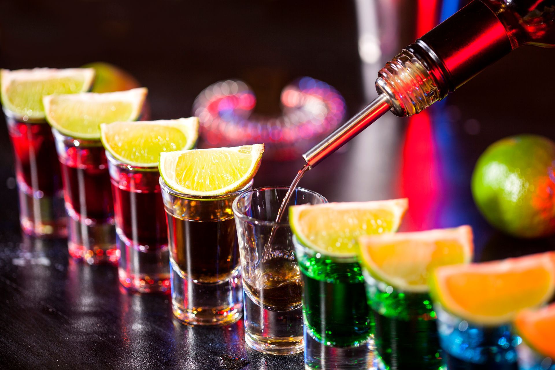 Row of colorful shots with lime wedges, being poured from a spout.