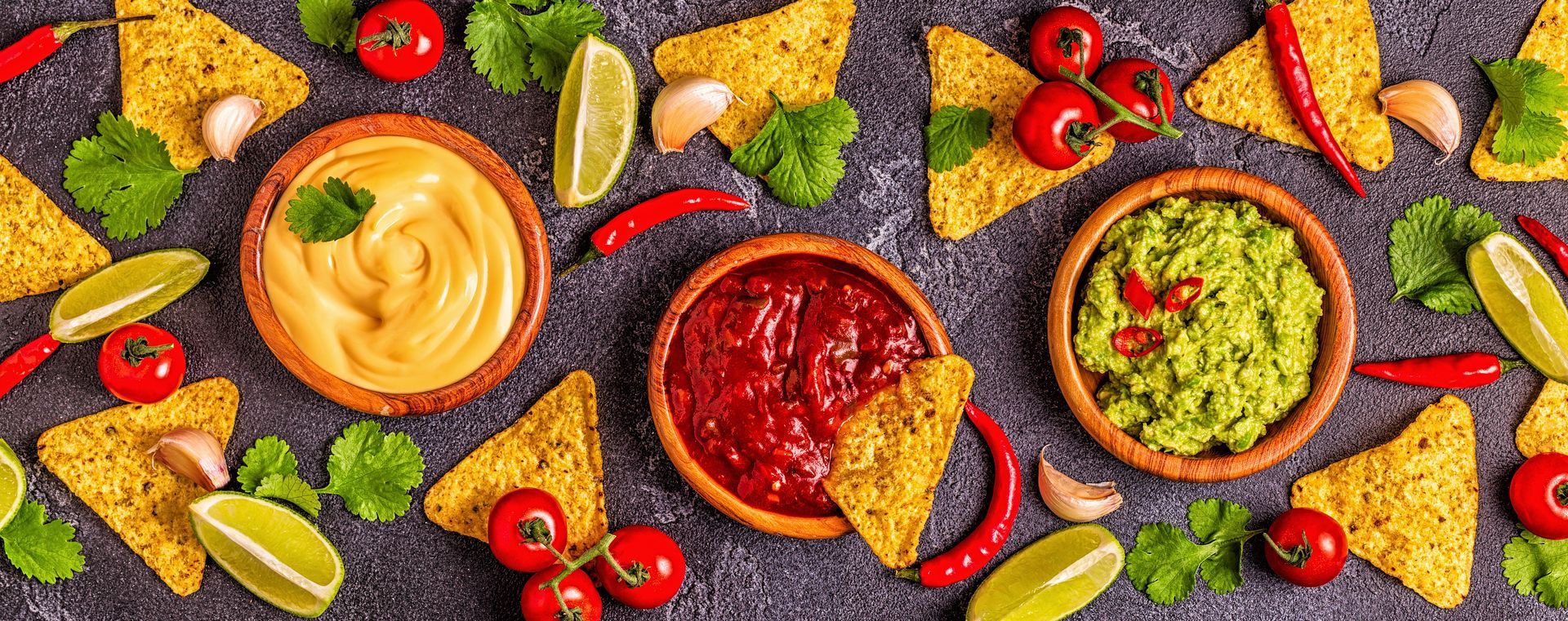 Overhead shot of tortilla chips, cheese dip, salsa, guacamole, tomatoes, peppers, cilantro, lime.