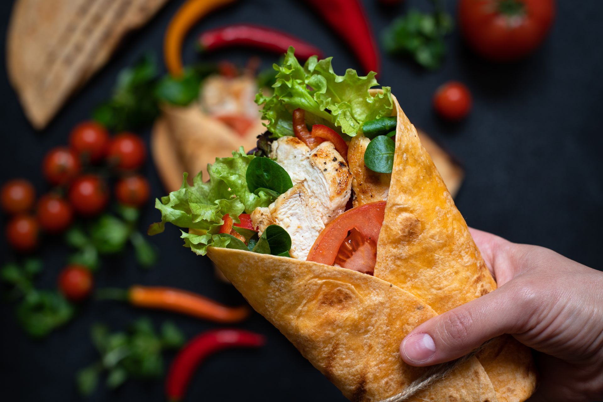 Hand holding a chicken wrap filled with lettuce, tomato, and vegetables, with blurred ingredients in the background.