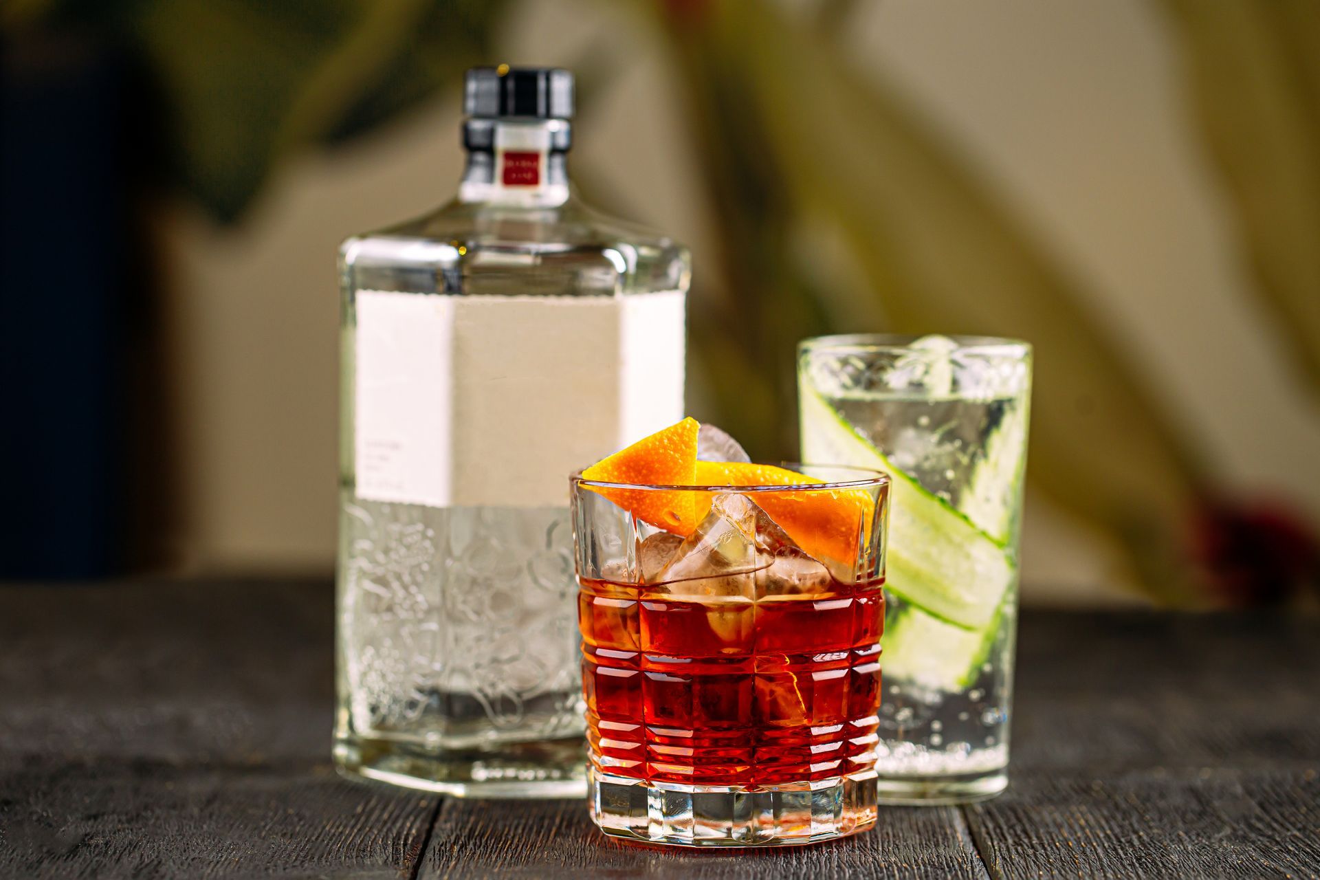 Bottle of gin with two cocktails on a dark wood surface. One red, one clear with cucumber slices.