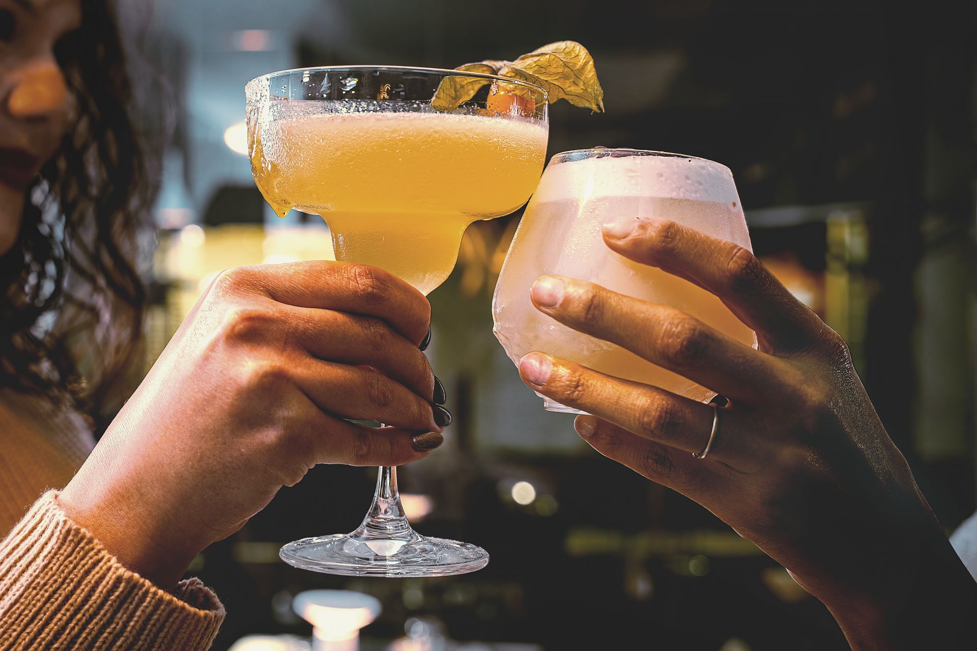 Two people toasting with cocktails; one in a margarita glass, other in a stemless glass.