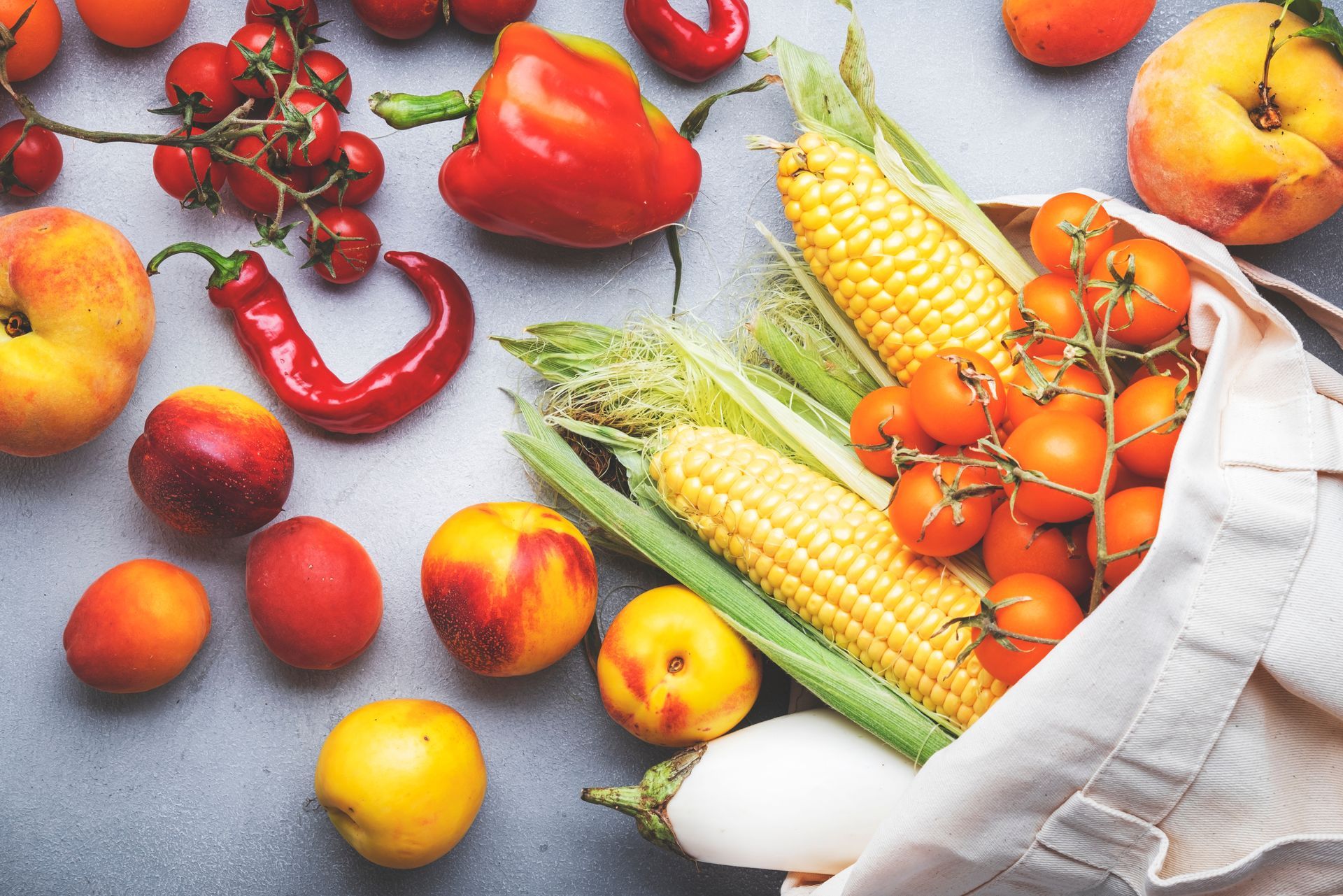 Fresh produce, including corn, tomatoes, peppers, peaches, and nectarines, spills from a canvas bag onto a gray surface.