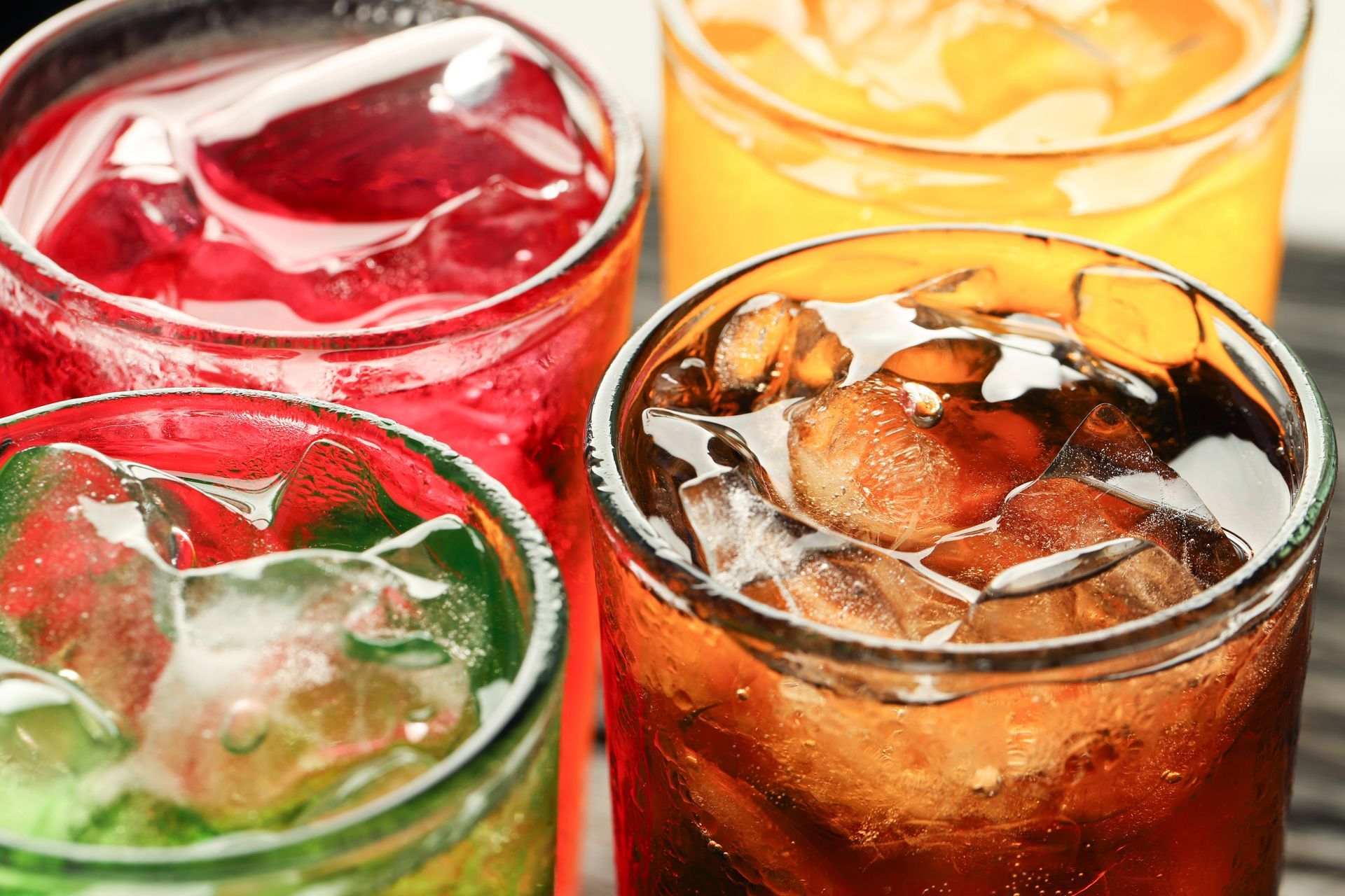 Four glasses of soda with ice; red, green, orange, and brown beverages, close-up.