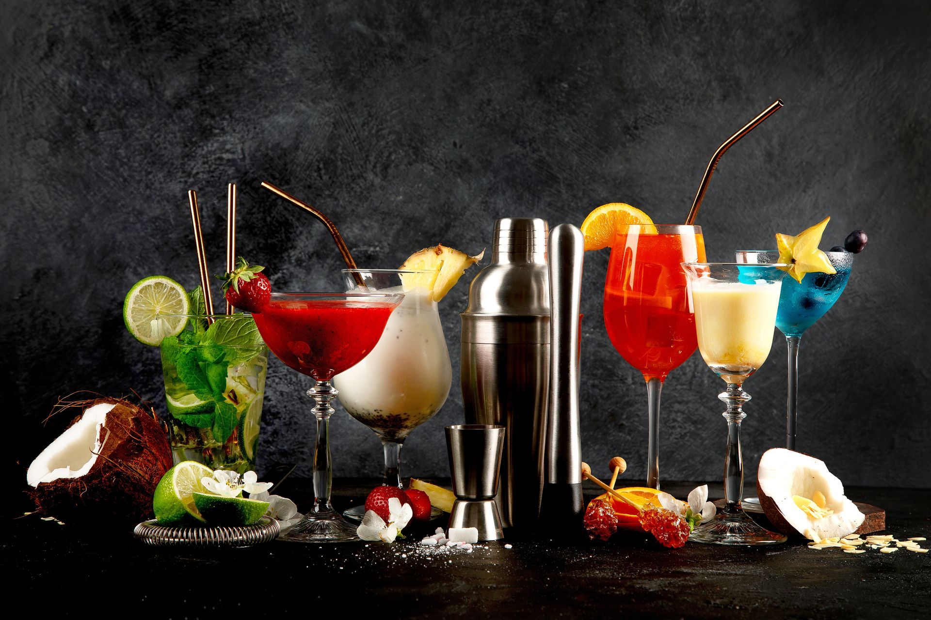 Cocktails in various glasses with garnishes, straws, and a shaker set against a dark background.
