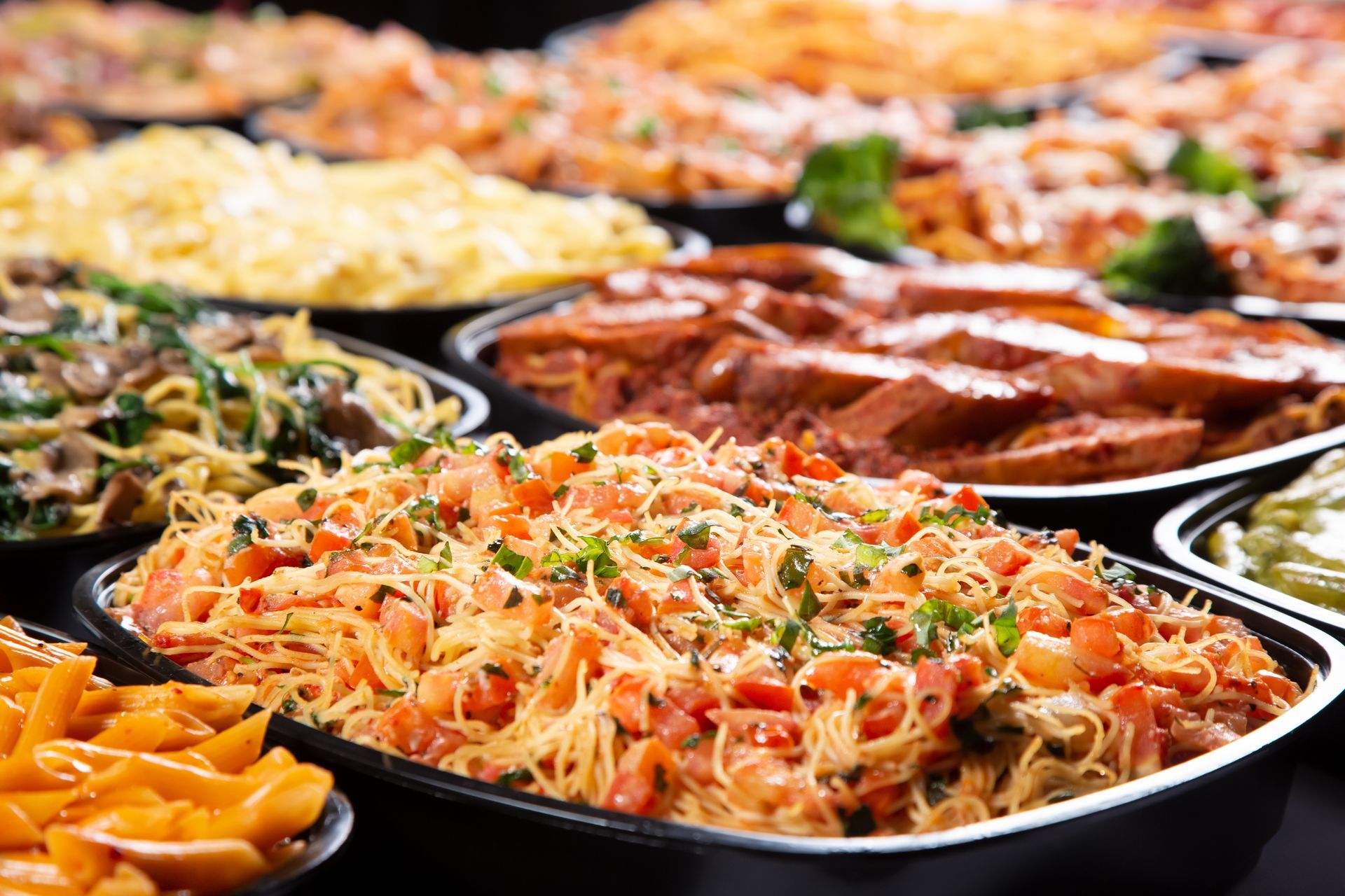 Various pasta dishes in black serving trays, brightly lit, with varied sauces and toppings.