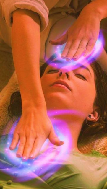 Manos sosteniendo un cuarzo rosa, rodeado de cristales, cartas del tarot y un manojo de salvia sobre una superficie oscura.