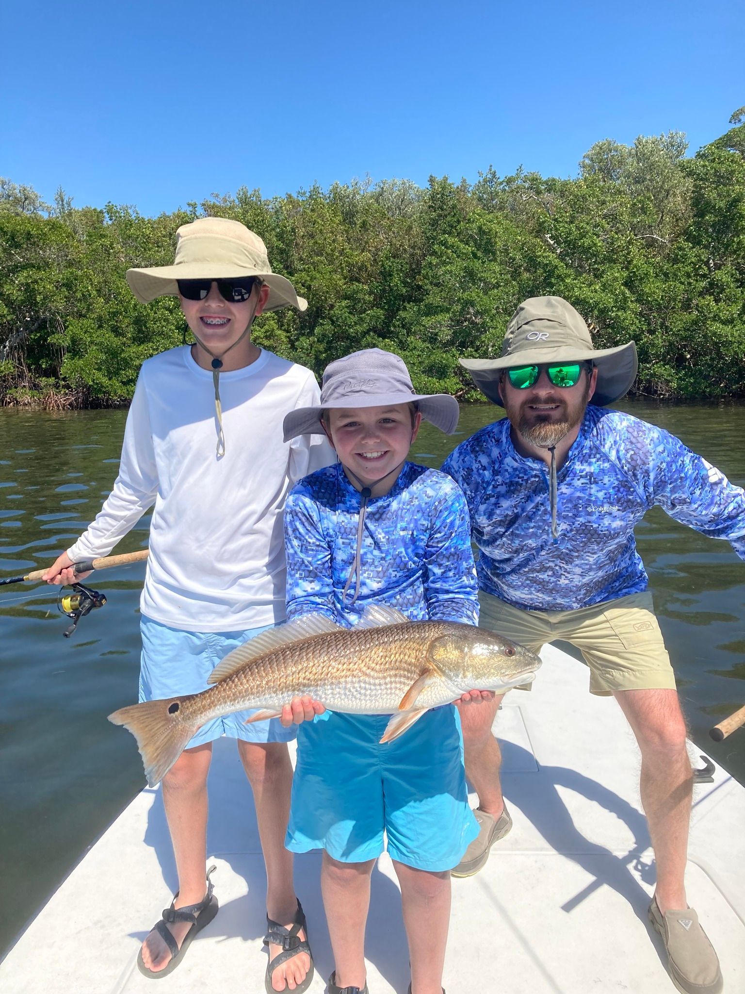 Three people on a boat holding a large reddish fish. The boy in the middle holds the fish, smiling, flanked by two men. They are in a sunny, mangrove-lined waterway.