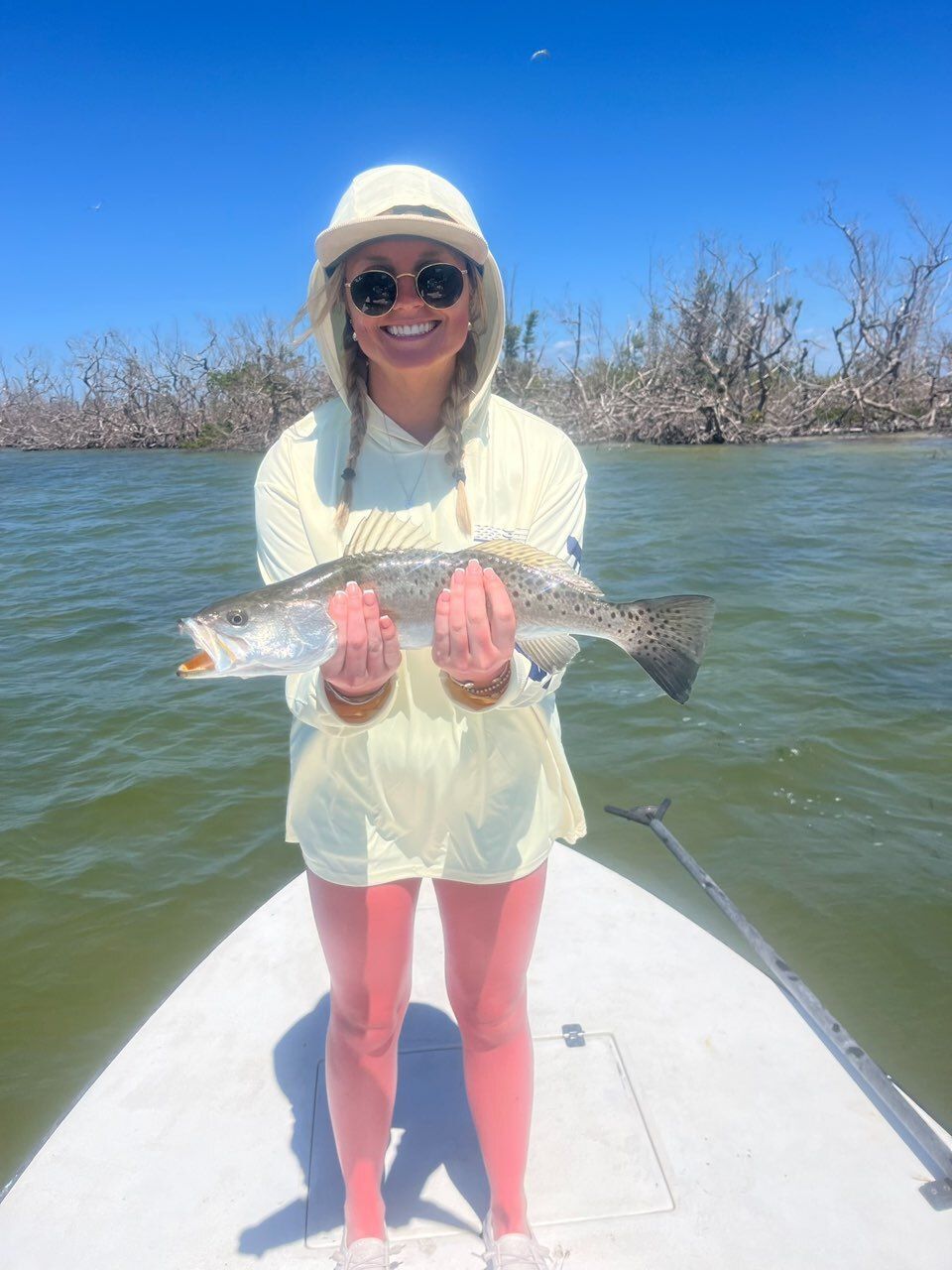 Woman on a paddleboard holds a speckled trout she caught in the water. She wears a sun hat, sunglasses, and light-colored clothing.