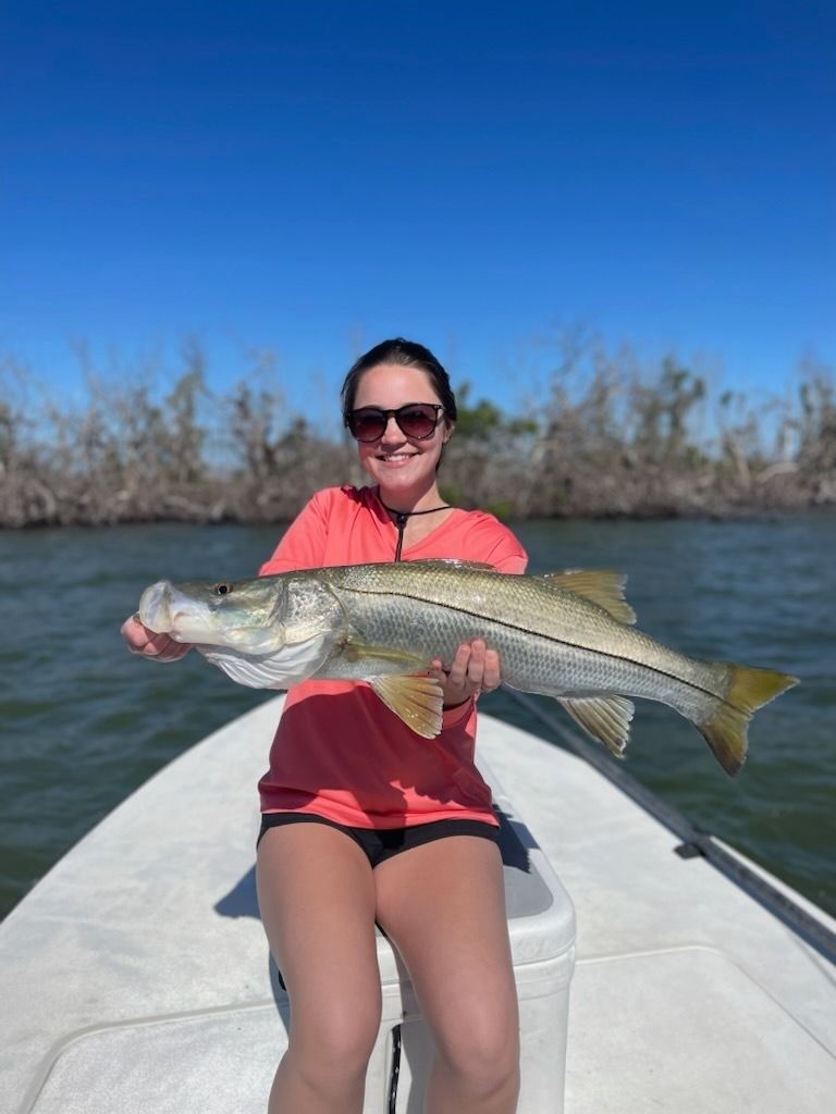 Woman on a boat holding a large silver fish, smiling under a bright blue sky. The background shows trees.
