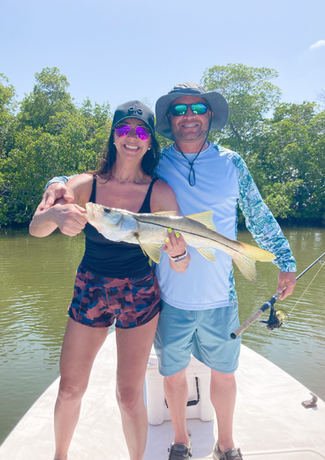 Man and woman on a boat smiling, holding up a fish they caught. They are in front of green trees on a sunny day.