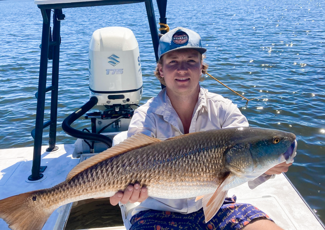 Young man in a cap on a boat, holding a large redfish. Sunlight reflects off the water and boat.
