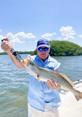 Man holding a speckled trout on a sunny day by the water. He wears a blue shirt, hat, and sunglasses.