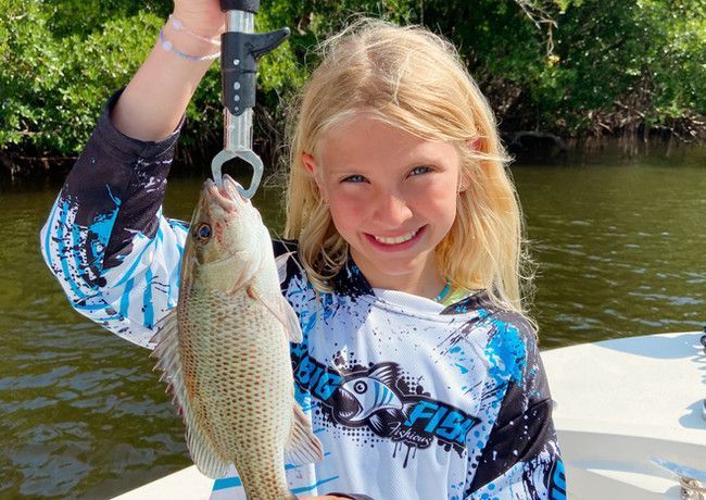 Girl smiling, holding a fish suspended by a hook, on a boat in a sunny outdoor setting.