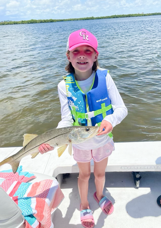 Girl in pink hat and life vest holds a fish on a boat, smiling at the camera. Sunny day on the water.