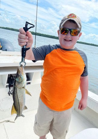 Young boy smiling, holding a fish with a scale on a boat, wearing sunglasses, an orange and gray shirt, and a baseball cap.