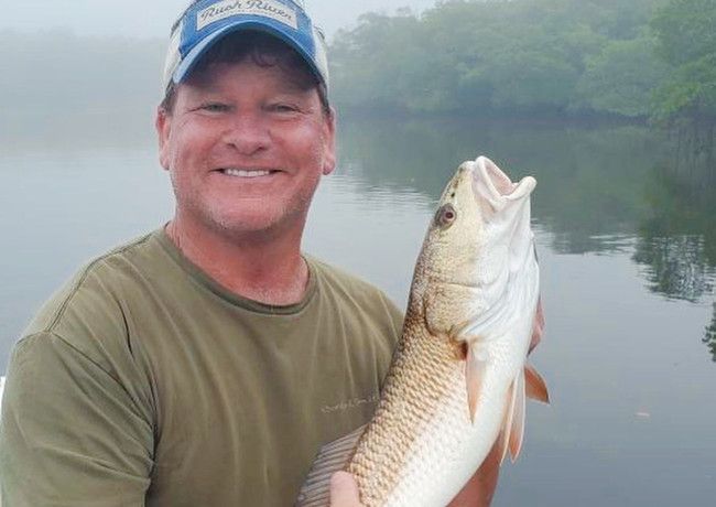 Man smiling, holding a large redfish on a boat, in a foggy waterway. He's wearing a hat and green shirt.