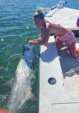 Woman on a boat in shorts and bikini top kneeling to release a large tarpon back into the water. Blue water, sunny day.