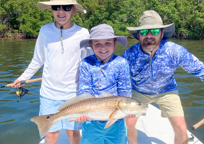 Three people on a boat, smiling and holding a redfish. They are on the water in a sunny location, with trees in the background.