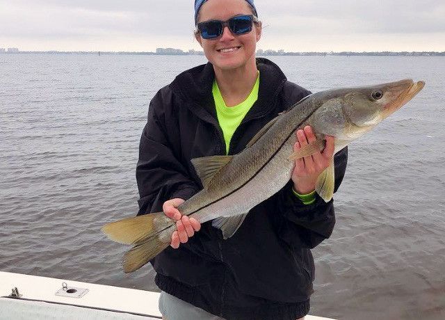 Woman smiling, holding a large silver fish on a boat. She wears sunglasses, a black jacket, and a bright green shirt.