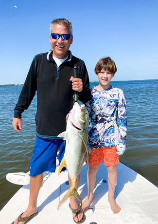 Man and boy on a boat display a large yellow fish they caught. They smile in bright sunlight with water in the background.