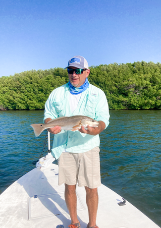 Man on a boat holding a fish. He is smiling, with trees in the background and blue water.