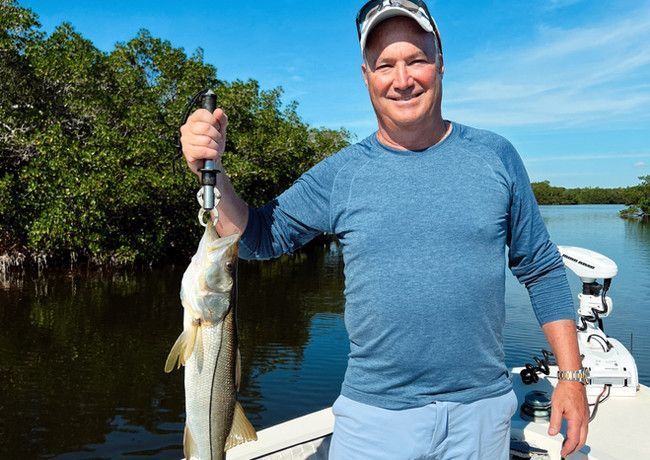 Man in a boat holds up a fish he caught, smiling against a backdrop of blue sky and mangrove trees.