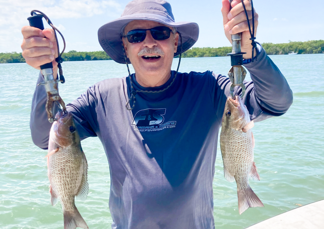 Man holding two fish he caught, smiling, wearing sunglasses, a hat, and a long-sleeved shirt, on a boat in the water.