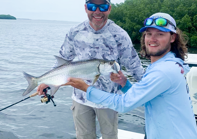 Two men on a boat, one holding a silver tarpon fish. The water is gray; they are smiling.