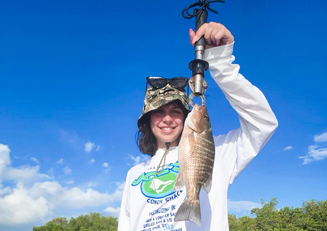 Person smiles, holding up a fish with a scale in bright sunshine. They wear a white shirt, camo hat, and sunglasses with blue sky and trees in the background.