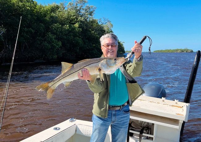 Man on a boat holds up a large fish he caught, smiling with joy against a backdrop of water and trees.