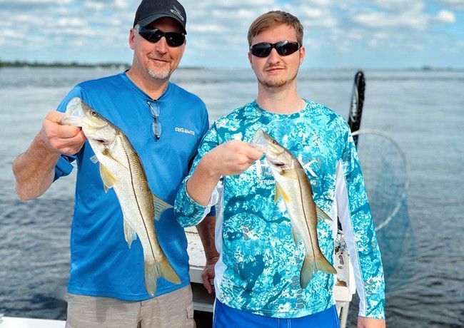 Two men on a boat holding up snook fish. One man wears blue, the other turquoise. The setting is a body of water.