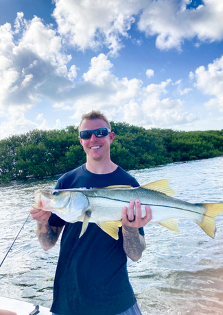 Man smiling, holding a large fish with yellow tail, on a boat near mangrove trees under a cloudy sky.