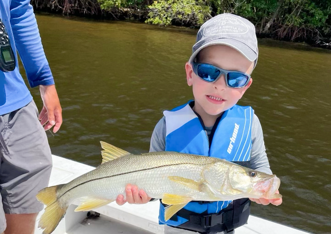 Young boy in sunglasses and a life vest holding a fish on a boat. The fish is silvery with yellow fins.