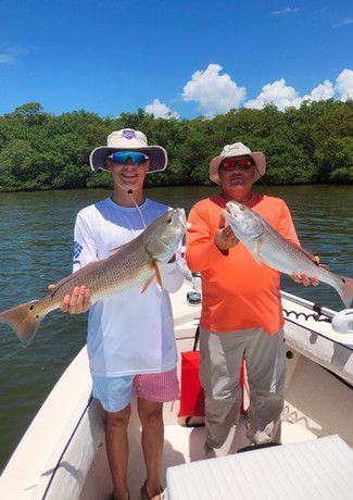 Two people on a boat hold up large redfish. The man on the left smiles, the other man is neutral. Sunny day, green trees in the background.