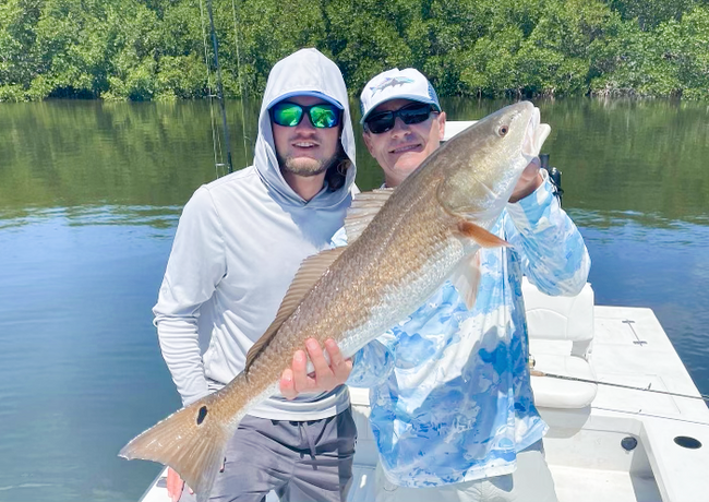 Two men on a boat holding a large redfish. Both are smiling. They are in front of a body of water with trees in the background.