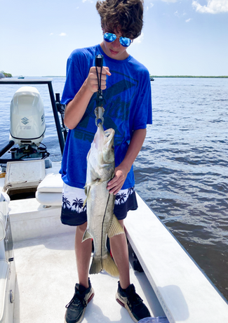 Teenager on a boat holds up a fish he caught, smiling, near a body of water. Blue sky and water, sunny day.