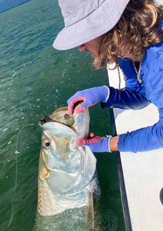 Person in hat and blue sleeves holding a large, silvery tarpon in the water, near a boat.