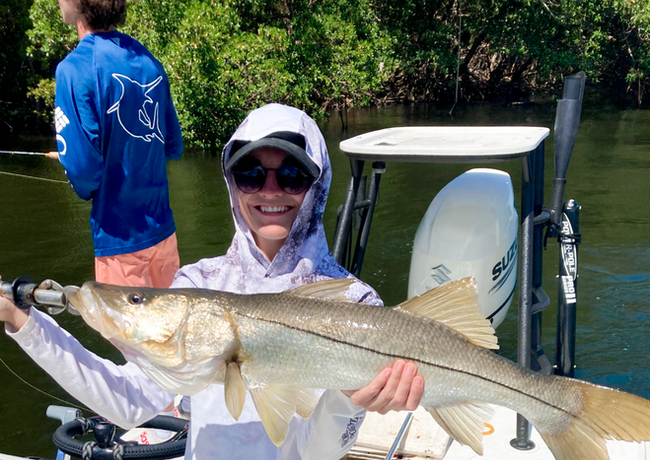 Woman smiling, holding a large silver snook fish on a boat. Wearing sunglasses, a hat, and long-sleeved shirt; another person in the background.