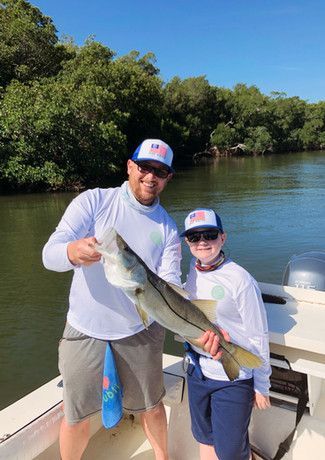 A man and child on a boat holding a large fish, likely a snook, with a lush green background. They wear matching white shirts and hats.