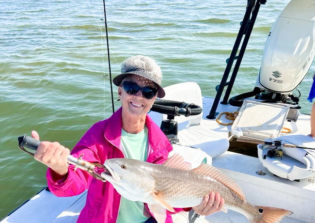 Woman on a boat smiling, holding a large redfish. She wears sunglasses, a hat, and pink jacket; water in the background.