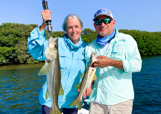 Two people on a boat fishing, holding up snook. They're smiling in front of mangroves and blue water.